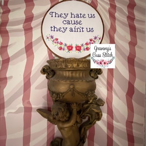K&ouml;nnte beinhalten: Ein Kreuzstich-Stickrahmen mit dem Text "They hate us cause they ain't us" und floralen Akzenten. Der Rahmen wird auf einer goldfarbenen, von einem Cherub getragenen Urne vor einem rosa-wei&szlig; gestreiften Hintergrund pr&auml;sentiert.