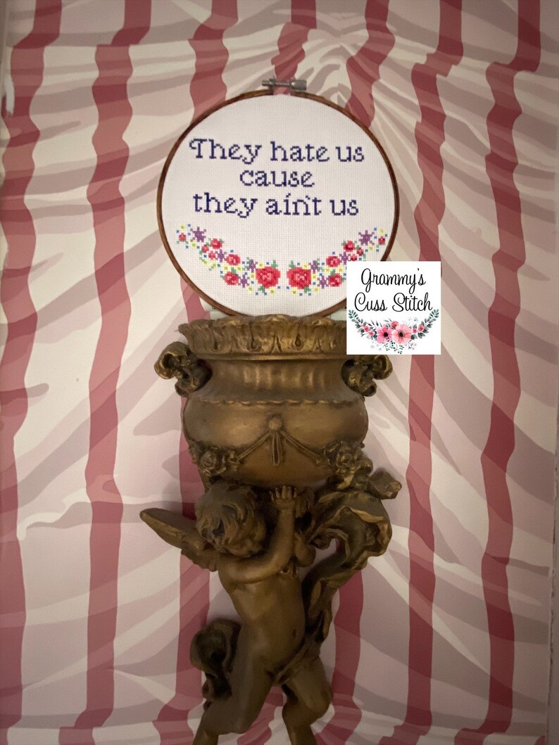 K&ouml;nnte beinhalten: Ein Kreuzstich-Stickrahmen mit dem Text "They hate us cause they ain't us" und floralen Akzenten. Der Rahmen wird auf einer goldfarbenen, von einem Cherub getragenen Urne vor einem rosa-wei&szlig; gestreiften Hintergrund pr&auml;sentiert.