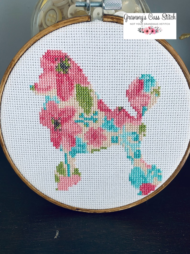 May include: Cross-stitch embroidery of a poodle dog filled with pink, green, and blue floral patterns. The embroidery is set in a wooden hoop. The text "Grammy's Cuss Stitch" is in the upper right corner.