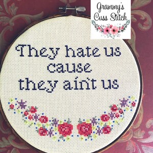 K&ouml;nnte beinhalten: Ein Kreuzstich-Stickrahmen mit dem Text "They hate us cause they ain't us" in dunkelblauem Garn. Das Design umfasst einen floralen Rand mit roten, rosa und lila Blumen. Der Rahmen ist vor einem burgunderfarbenen Hintergrund platziert.