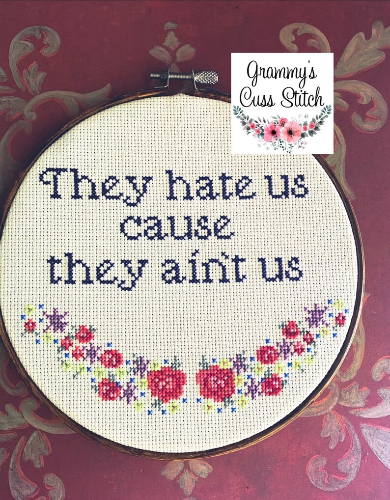 K&ouml;nnte beinhalten: Ein Kreuzstich-Stickrahmen mit dem Text "They hate us cause they ain't us" in dunkelblauem Garn. Das Design umfasst einen floralen Rand mit roten, rosa und lila Blumen. Der Rahmen ist vor einem burgunderfarbenen Hintergrund platziert.
