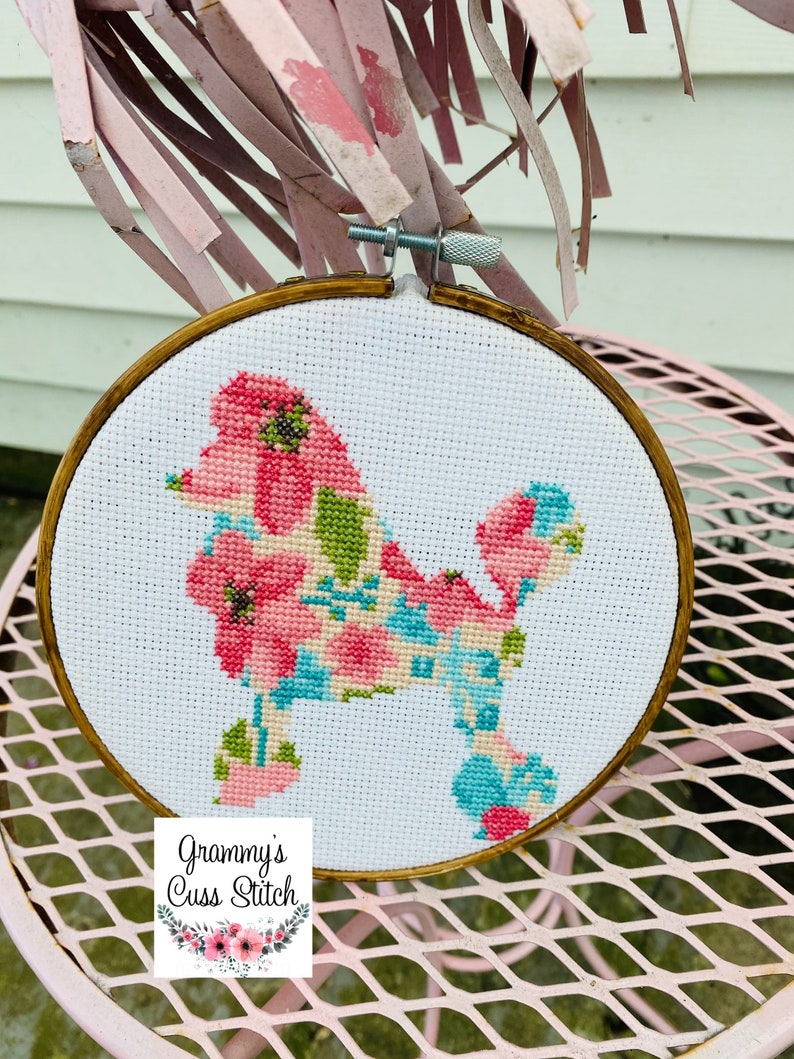 May include: A cross-stitch embroidery hoop featuring a poodle design. The poodle is filled with pink, blue, and green floral patterns. The hoop is set against a white background and framed in a wooden hoop. The text "Grammy's Cuss Stitch" is at the bottom.
