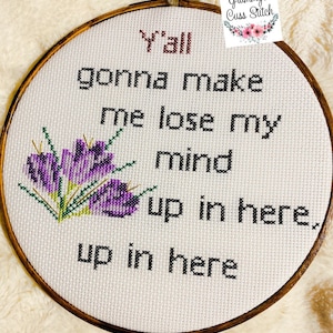 May include: A cross-stitch embroidery hoop with the text "Y'all gonna make me lose my mind up in here, up in here." The text is in black, with "Y'all" in red. Purple flowers are stitched on the left. The hoop is wooden.