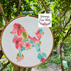 May include: Cross-stitch embroidery of a poodle silhouette filled with pink, blue, and green floral patterns. The embroidery is set in a wooden hoop and includes the text "Grammy's Cuss Stitch". The background is a natural outdoor setting with green foliage.