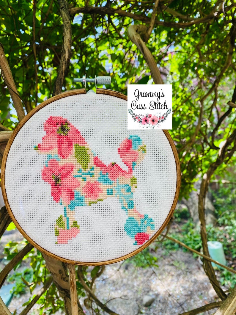 May include: Cross-stitch embroidery of a poodle silhouette filled with pink, blue, and green floral patterns. The embroidery is set in a wooden hoop and includes the text "Grammy's Cuss Stitch". The background is a natural outdoor setting with green foliage.