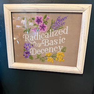 May include: A framed cross-stitch embroidery on tan fabric featuring the white text "Radicalized by Basic Decency" surrounded by embroidered purple, yellow, and white flowers. The piece is enclosed in a white wooden frame with a visible wood grain.