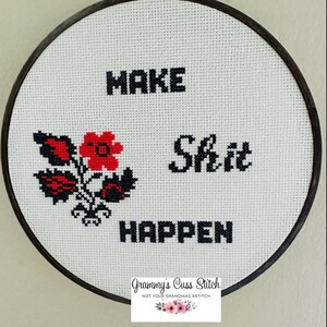 K&ouml;nnte beinhalten: Kreuzstich-Stickerei in einem Holzreifen. Der wei&szlig;e Stoff zeigt die Worte "MAKE Shit HAPPEN" in schwarzen Blockbuchstaben, mit einem rot-schwarzen Blumendesign. In der unteren rechten Ecke befindet sich ein Etikett mit der Aufschrift "Grammy's Cuss Stitch".