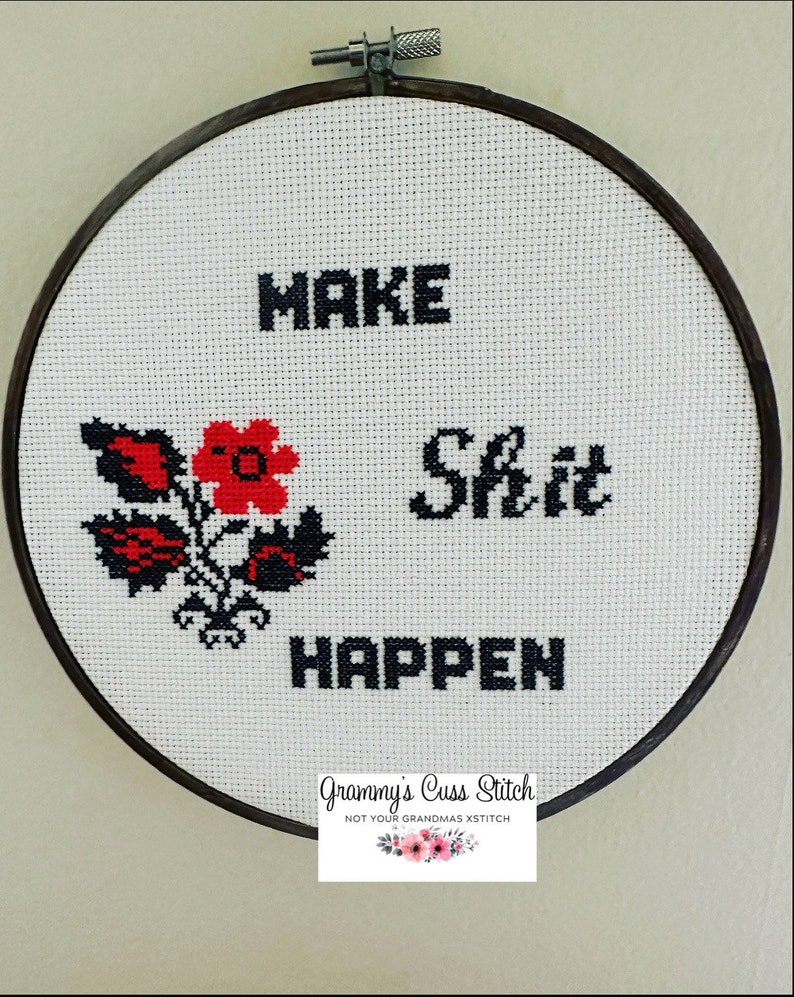 K&ouml;nnte beinhalten: Kreuzstich-Stickerei in einem Holzreifen. Der wei&szlig;e Stoff zeigt die Worte "MAKE Shit HAPPEN" in schwarzen Blockbuchstaben, mit einem rot-schwarzen Blumendesign. In der unteren rechten Ecke befindet sich ein Etikett mit der Aufschrift "Grammy's Cuss Stitch".