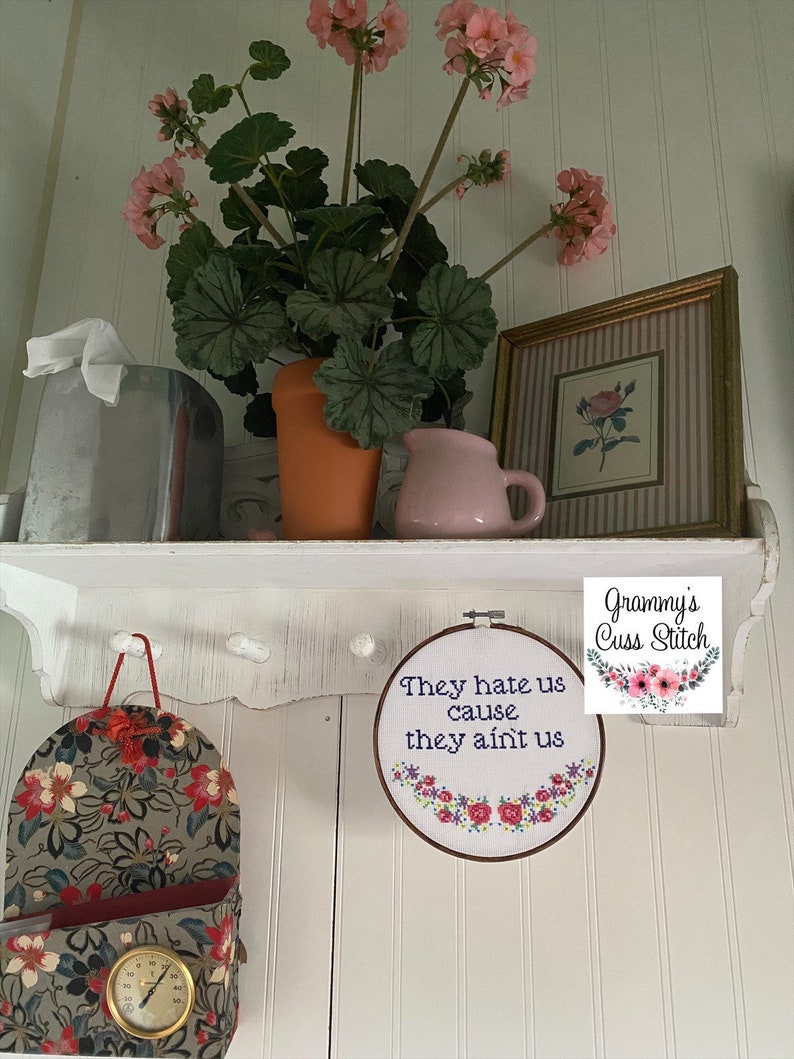 K&ouml;nnte beinhalten: Ein wei&szlig;es Regal zeigt ein gerahmtes Bild, eine rosa Kanne und einen Terrakottatopf mit rosa Blumen. Darunter h&auml;ngt ein floraler Stickrahmen mit dem Text "They hate us cause they ain't us". Ein floraler Stofforganizer ist ebenfalls sichtbar.