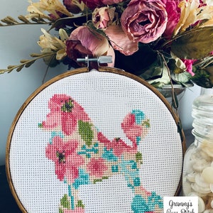 May include: A cross-stitch embroidery of a poodle silhouette filled with pink, blue, and green floral patterns. The embroidery is set in a round wooden hoop. The image includes the text "Grammy's Cuss Stitch".