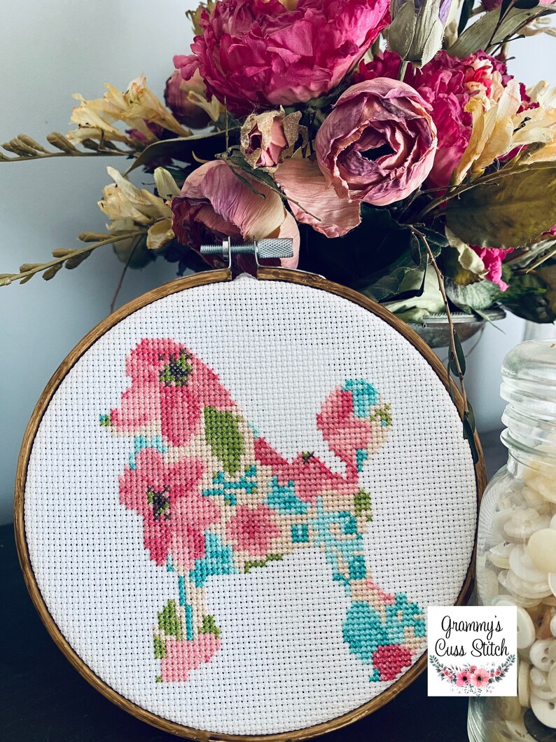 May include: A cross-stitch embroidery of a poodle silhouette filled with pink, blue, and green floral patterns. The embroidery is set in a round wooden hoop. The image includes the text "Grammy's Cuss Stitch".