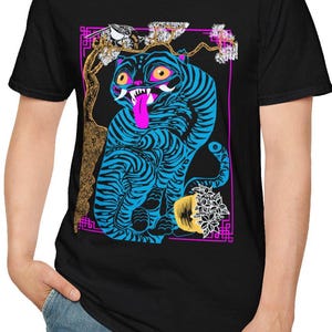 May include: Black t-shirt featuring a blue tiger design with black stripes, a pink tongue, and yellow eyes. The graphic includes a bird, flowers, and a gold tree branch, all within a pink and purple border.