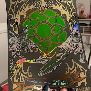 May include: A black art print with gold and green accents. The print features a deer-like creature and a hand reaching out, with the text "Perhaps now, we can both be human." The artwork has a stained glass window design.
