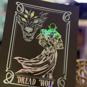 Puede incluir: Una impresión holográfica negra y plateada con una cabeza de lobo y una figura con una capa. El texto "Dread Wolf" está en la parte inferior de la impresión.