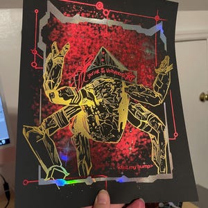 Può includere: Stampa artistica nera con una figura robotica dorata e nera su sfondo rosso e argento. Il testo "SAVIOR of the UNIVERSE" e "<fist.my bump>" è visibile, con un effetto olografico.