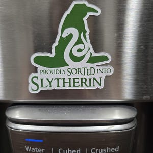 May include: A green and white sticker featuring the Sorting Hat and a snake, with the text "PROUDLY SORTED INTO SLYTHERIN." Below, a water dispenser with options for water, cubed ice, and crushed ice.