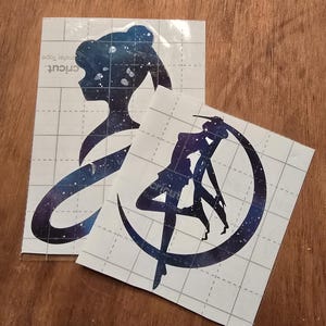 May include: Two vinyl decals featuring a silhouette of a character with a galaxy design. One shows a profile, the other a figure within a crescent moon. The decals are on a grid-patterned backing with the word "cricut" visible.