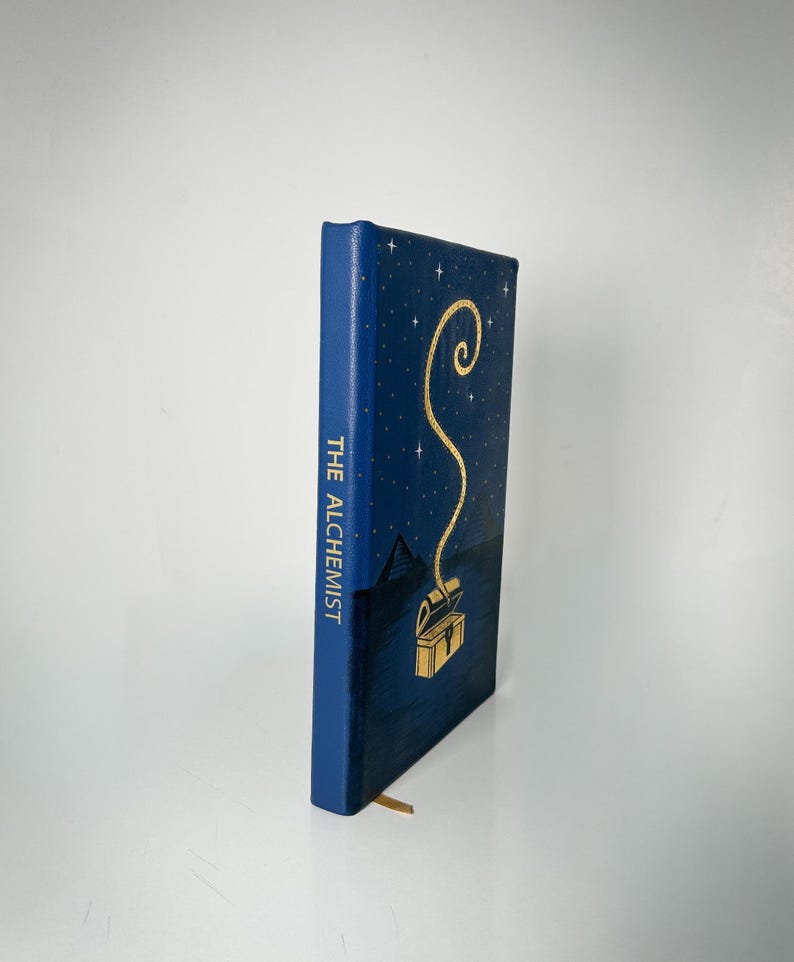 May include: A hardback book titled "The Alchemist" with a dark blue cover. It features a gold illustration of a treasure chest and a winding line. The spine is dark blue with the title in gold. A gold bookmark is visible.