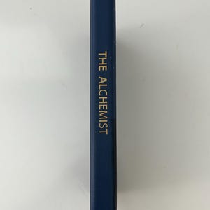 May include: A navy blue book with the title "THE ALCHEMIST" in gold lettering. The book has a leather bookmark. The book is on a white surface.