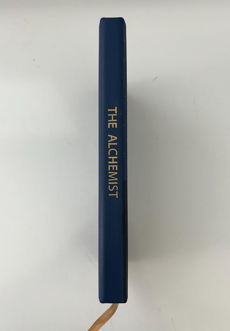 May include: A navy blue book with the title "THE ALCHEMIST" in gold lettering. The book has a leather bookmark. The book is on a white surface.