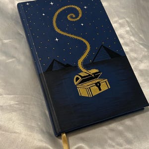May include: A navy blue journal with a gold treasure chest design. The cover features a starry night sky with pyramids and a golden spiral emanating from the chest. A gold ribbon bookmark is visible.