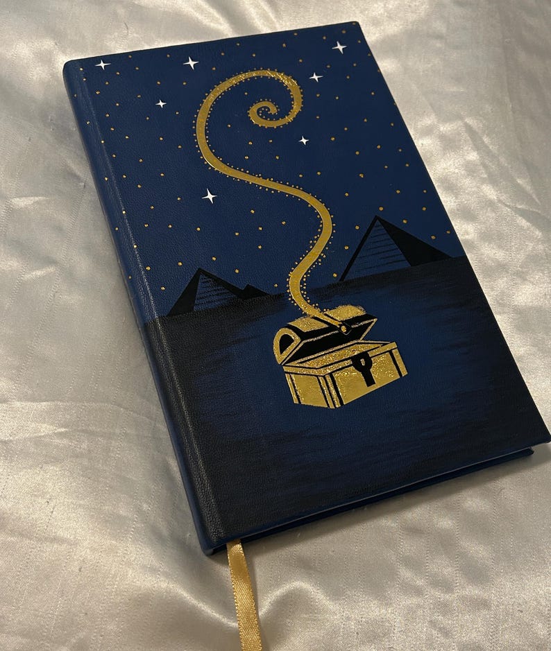 May include: A navy blue journal with a gold treasure chest design. The cover features a starry night sky with pyramids and a golden spiral emanating from the chest. A gold ribbon bookmark is visible.