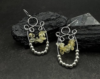 May include: A pair of handcrafted silver earrings. Each earring features a wire design with a cluster of yellow-green gemstones and a row of small silver beads. The earrings have a hook closure.