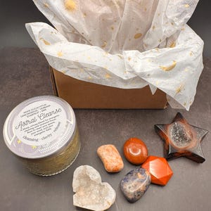 May include: A gift set featuring a brown box lined with tissue paper printed with gold celestial designs. The set includes a candle labeled "Astral Cleanse" and various crystals in shades of orange, red, and blue, along with a star-shaped geode.