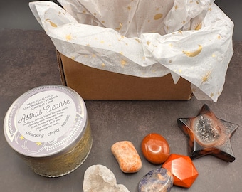 May include: A gift set featuring a brown box lined with tissue paper printed with gold celestial designs. The set includes a candle labeled "Astral Cleanse" and various crystals in shades of orange, red, and blue, along with a star-shaped geode.