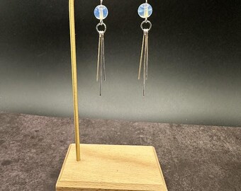 May include: A pair of dangle earrings displayed on a gold-colored stand. Each earring features a light blue, round bead, a small silver ring, and three silver, linear dangles. The earrings are set against a dark background.