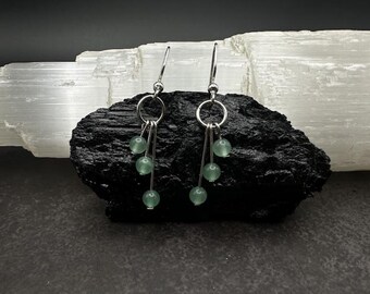 May include: A pair of dangle earrings with silver-tone hooks and three light green beads suspended from each earring. The earrings are displayed on a dark, textured surface with a white, crystalline background.