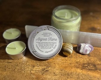 May include: An assortment of spiritual items on a wooden surface. The items include a candle in a glass jar, two small tea light candles, a silver tin with the text "Aligned Flame", two stones, and a long, white crystal. The tin's text includes "Frankincense Rosemary Sage Bergamot".
