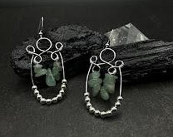 May include: A pair of silver wire earrings with a unique design. Each earring features a wire frame with a cluster of small, green stones and a row of silver beads. The earrings have a hook closure.