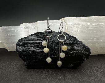 May include: A pair of silver earrings with a dangling design. Each earring features a circular top with three strands, each holding a small, round, off-white bead. The earrings are displayed on a dark, textured surface.