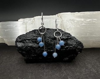 May include: A pair of silver-toned dangle earrings. Each earring features a circular top with three strands of small, light blue beads. The earrings are displayed on a black, textured surface against a white background.