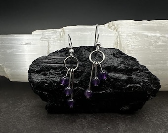May include: A pair of dangling earrings with silver-colored hooks and circular accents. Each earring features multiple strands of small, round, deep purple beads. The earrings are displayed on a dark, textured surface.
