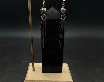 May include: A pair of dangle earrings with a starburst design. The earrings feature a gold-colored star at the top, a silver ring, and multiple silver-colored, thin, dangling bars. The earrings are displayed on a black rectangular stand with a wooden base.