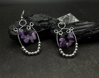 May include: A pair of silver-toned wire earrings. Each earring features a curved wire frame with a cluster of purple amethyst stones and a row of small silver beads. The earrings have a hook closure.