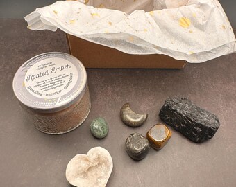 May include: A gift box with tissue paper featuring gold celestial designs. The box contains a candle labeled "Rooted Ember" and several crystals, including a heart-shaped stone, a moon-shaped stone, and a black tourmaline.