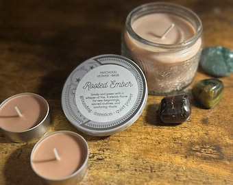 May include: A collection of candles and crystals. The image features a large jar candle and two small tea light candles, all with a light brown wax. A round container with the text "Rooted Ember" is also present, along with several polished stones.