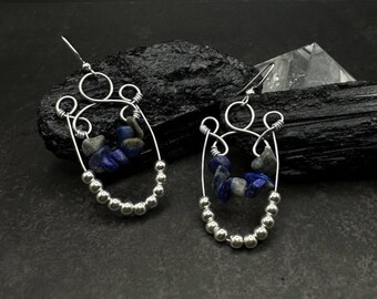 May include: A pair of handcrafted silver earrings. Each earring features a wire design with a series of small silver beads along the bottom and a cluster of blue and gray gemstones. The earrings have a hook closure.