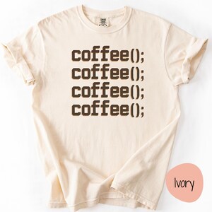 May include: Ivory-coloured t-shirt featuring the text "coffee();" repeated four times in a brown, blocky font. The shirt has short sleeves and a crew neck. The shirt is made of a soft, comfortable material.