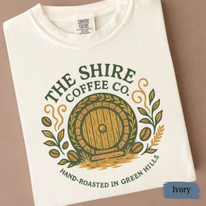 May include: Ivory-colored t-shirt featuring a circular design with the text "THE SHIRE COFFEE CO." and a graphic of a round wooden door. The design includes coffee beans and foliage, with the words "HAND-ROASTED IN GREEN HILLS" at the bottom.