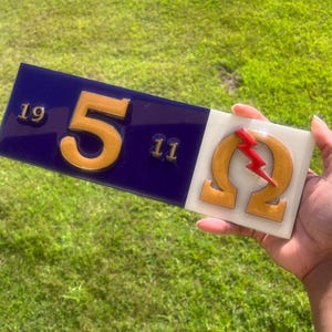 Op de afbeelding: Een tweedelige decoratieve plaat. De linkerkant is een donkerblauwe rechthoek met het gouden nummer "5", geflankeerd door de nummers "19" en "11". De rechterkant is een witte vierkant met een gouden Griekse Omega en een rode bliksem.