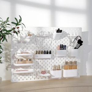 May include: A white pegboard organizer with various cosmetic and jewelry storage containers. The organizer holds lipsticks, brushes, perfumes, and other beauty products. The pegboard is mounted on a white wall, with a plant in the upper left corner.