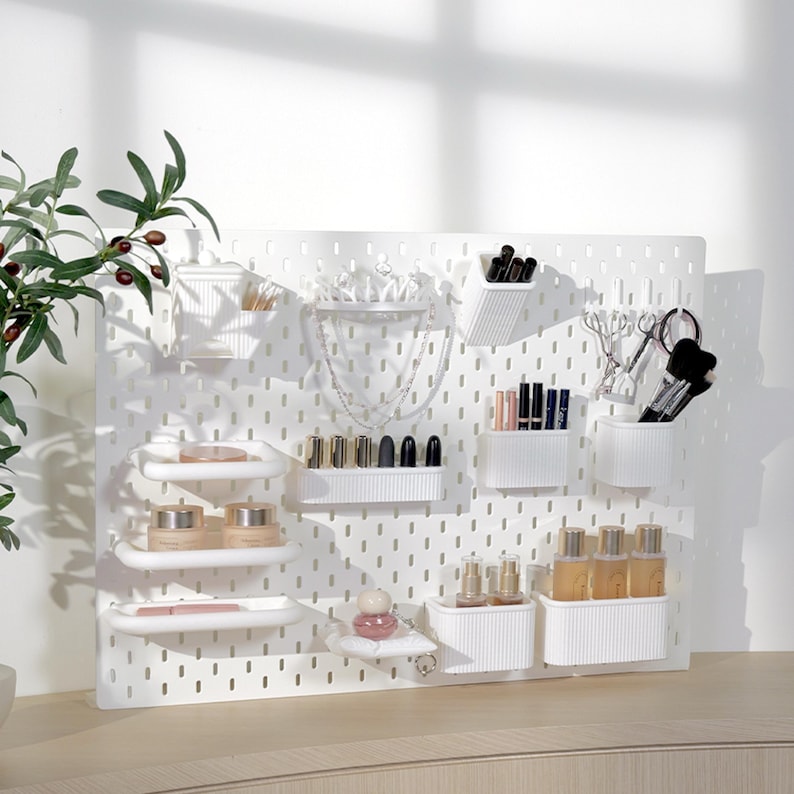 May include: A white pegboard organizer with various cosmetic and jewelry storage containers. The organizer holds lipsticks, brushes, perfumes, and other beauty products. The pegboard is mounted on a white wall, with a plant in the upper left corner.