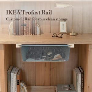 May include: A gray metal mesh basket suspended from a wooden shelf, labeled "IKEA Trofast Rail." The shelf is part of a wooden cabinet, with books and decorative items on the shelf. The basket is designed for storage.