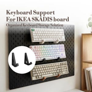 May include: A set of two black L-shaped keyboard support hooks designed for an IKEA SKADIS pegboard. The image shows three keyboards mounted on a black pegboard using these supports, with a circular inset highlighting the two black hooks.