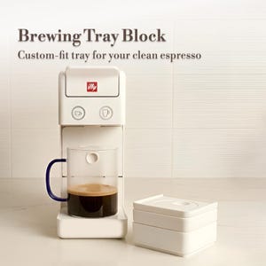 May include: A white espresso machine with the text "Brewing Tray Block" and "Custom-fit tray for your clean espresso." A clear glass mug with a blue handle is filled with coffee. A white tray block sits to the right.