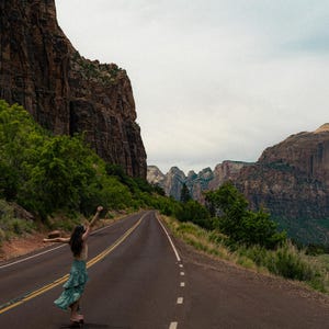 May include: A person standing on an asphalt road with arms raised, wearing a light-colored top and a green patterned skirt, set against a backdrop of towering red rock canyon walls, green trees, and a cloudy sky in a vast desert landscape.